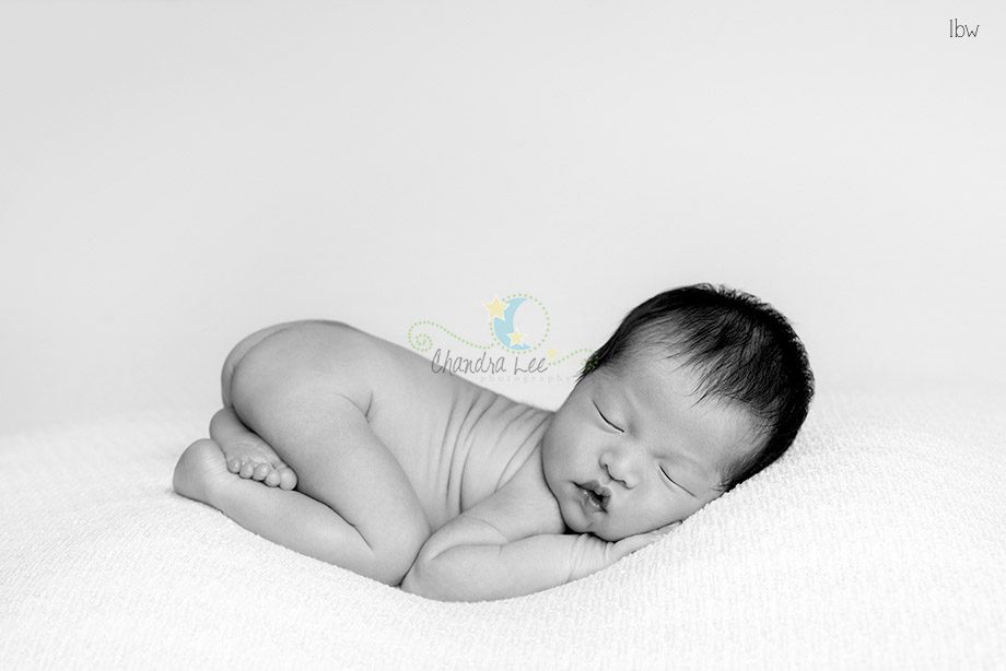 A newborn baby sleeping on a soft surface, curled up with arms and legs tucked close. The image is in black and white, showcasing the baby's peaceful expression. The background is a smooth, light color.