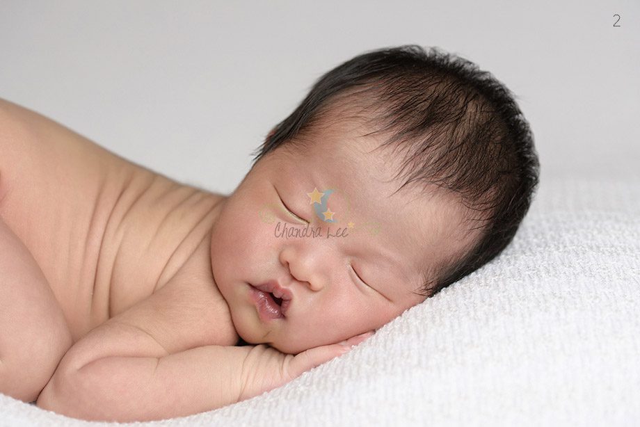 A sleeping newborn baby lying on a white blanket with arms tucked under the face. The baby has dark hair and a peaceful expression.
