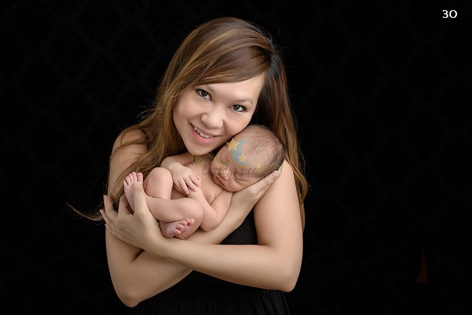 A woman wearing a black dress holds a swaddled newborn baby in her arms, smiling gently. The background is dark, emphasizing the subjects.
