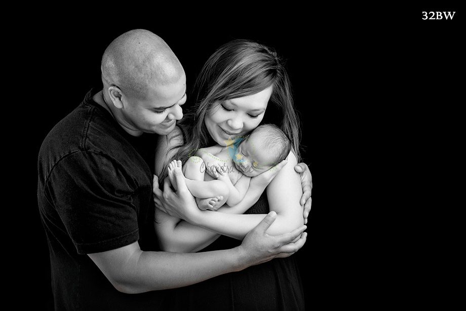 A couple holding a newborn baby in a close embrace against a black background. The mother is wearing a dark dress, and the father wears a black shirt. The baby is swaddled in a light fabric.