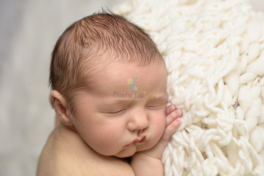 A close-up of a sleeping newborn baby with short hair resting on a soft, white, knitted blanket. The baby has a peaceful expression with hands tucked under the chin.