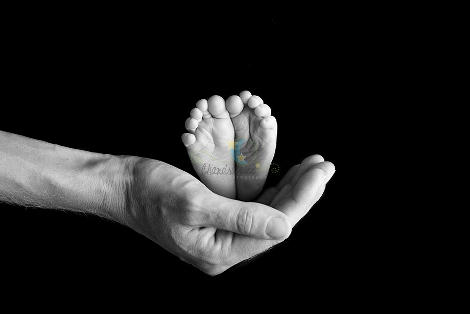 An adult hand cradling a small baby's foot against a black background. The baby's foot is facing upwards with visible toes.