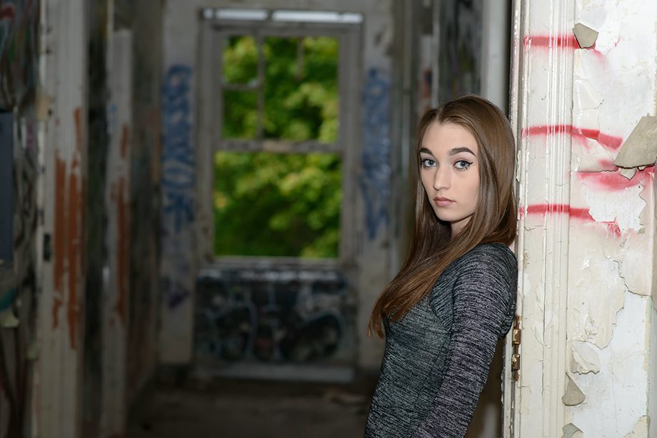 A young woman standing against the wall in a dimly lit hallway with graffiti on the walls. She has long brown hair and is looking directly at the camera. A window at the end of the hall shows green foliage outside.