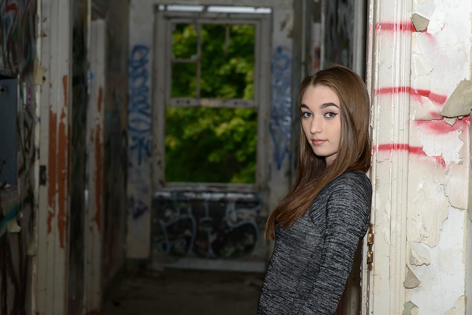 A young woman with long hair leans against a wall in a corridor with graffiti. Light enters through a window at the end of the hall, highlighting the greenery outside.